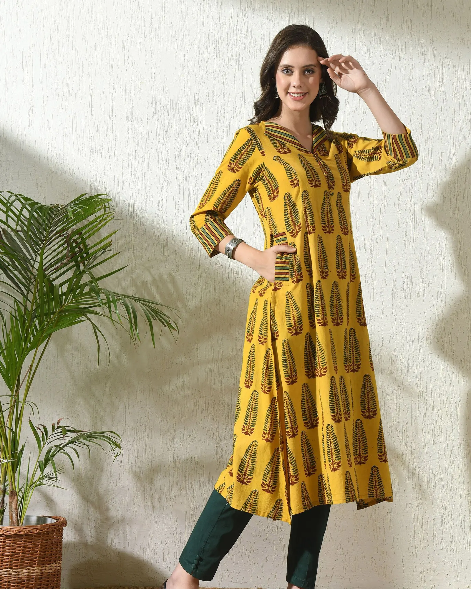 A woman in a mustard block-print kurta standing against a white wall beside a palm
